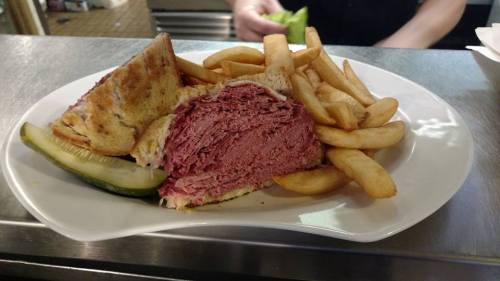 A plate with a large sandwich filled with corned beef, served with fries and a pickle on the side.