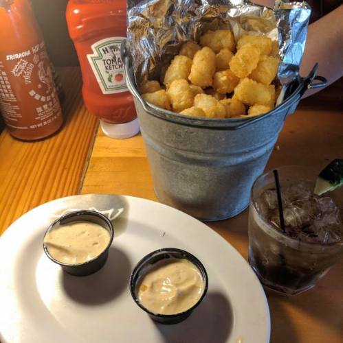 A bucket of tater tots next to two dipping sauces and a drink on a wooden table, with ketchup bottles in the background.