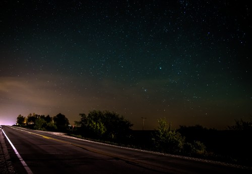 A dark road stretches into the distance under a starry night sky, with faint clouds and silhouettes of trees on the sides.