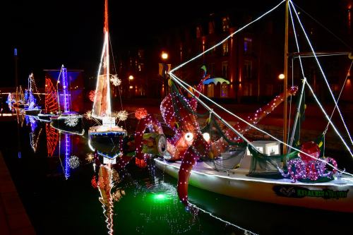 Colorful boats decorated with lights float on a canal at night, reflecting vibrant colors in the water.