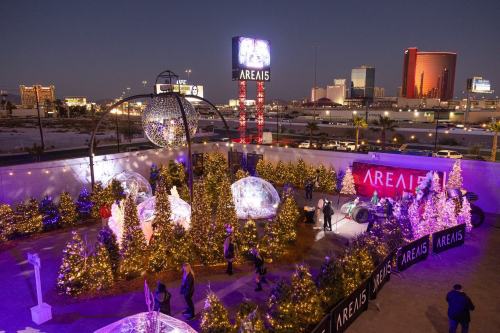 A festive outdoor scene at AREA15 with illuminated trees, decorations, and a large disco ball at dusk.