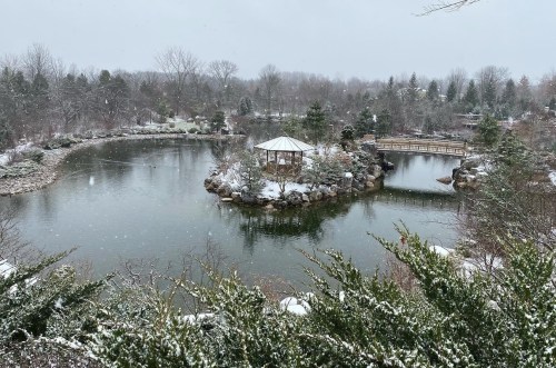 A serene winter scene of a pond surrounded by snow-covered trees and a gazebo, with light snowfall in the air.