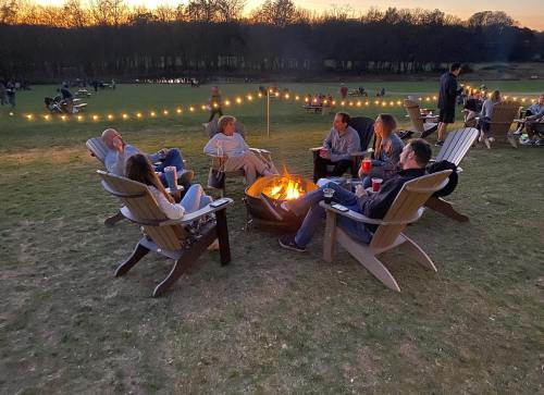 A group of people sitting around a fire pit at sunset, enjoying drinks and conversation in a grassy outdoor setting.