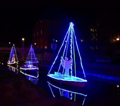 Two illuminated boats with colorful lights, one shaped like a tree, sailing on a dark canal at night.