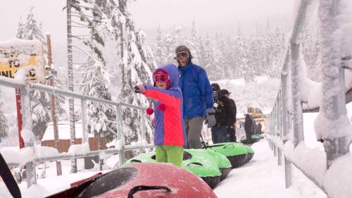A child in a colorful jacket and goggles points while waiting in line for snow tubing, with adults and snowy scenery around.
