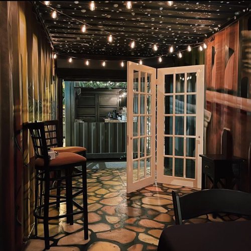 Cozy interior with string lights, an open door, and a bar stool, featuring a painted stone floor and colorful walls.