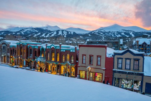 Snow-covered buildings line a street, with mountains in the background and a colorful sunset sky.