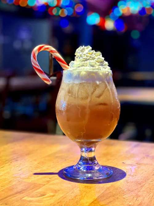 A festive drink in a glass, topped with whipped cream and a candy cane, set against a colorful, blurred background.