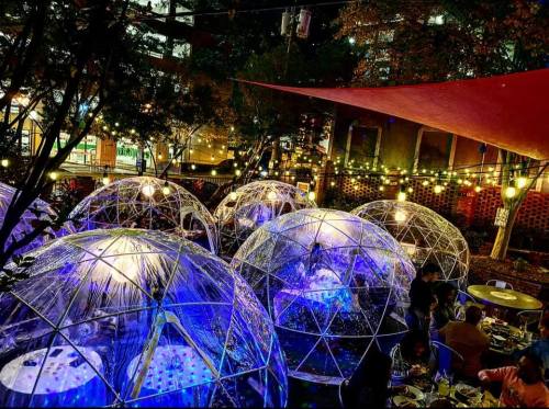 Colorful illuminated dining domes in a cozy outdoor setting, surrounded by trees and string lights at night.