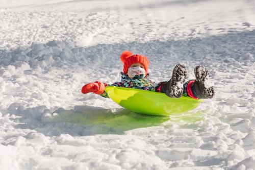 A child in a colorful winter outfit sleds down a snowy hill, wearing a bright orange hat and gloves.