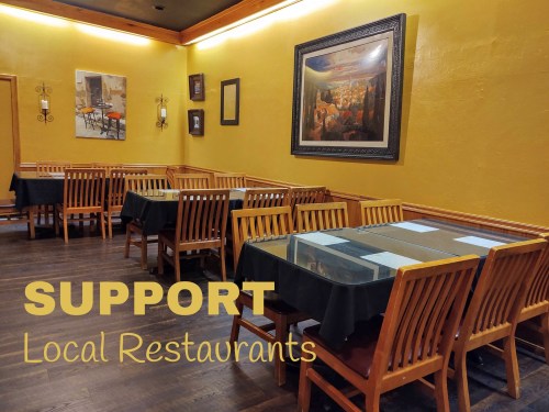 A cozy restaurant interior with wooden tables and chairs, featuring a warm yellow wall and a call to support local restaurants.