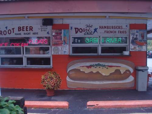 A colorful fast-food stand featuring neon signs for hot dogs, hamburgers, and root beer, with a painted hot dog on the wall.