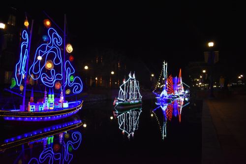 Colorful illuminated boats with swirling patterns and bright lights reflect on a calm waterway at night.