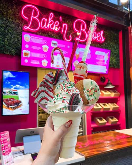 A hand holds a festive ice cream sundae topped with cookies, candy canes, and a Santa decoration in front of a colorful shop.
