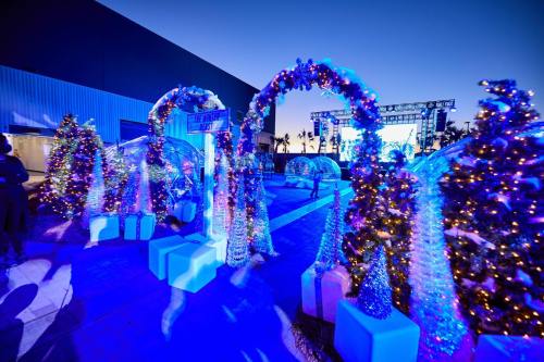 A festive outdoor scene with illuminated arches, decorated trees, and a stage in the background, all under a twilight sky.