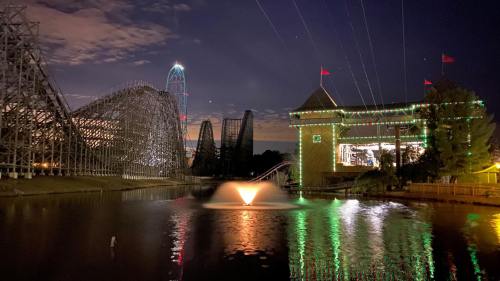 Nighttime view of a theme park with illuminated roller coasters and a fountain reflecting colorful lights in the water.