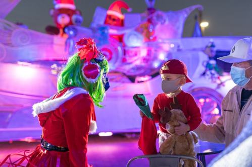 A child in a red cap and mask holds a stuffed reindeer, interacting with a performer in a festive costume.