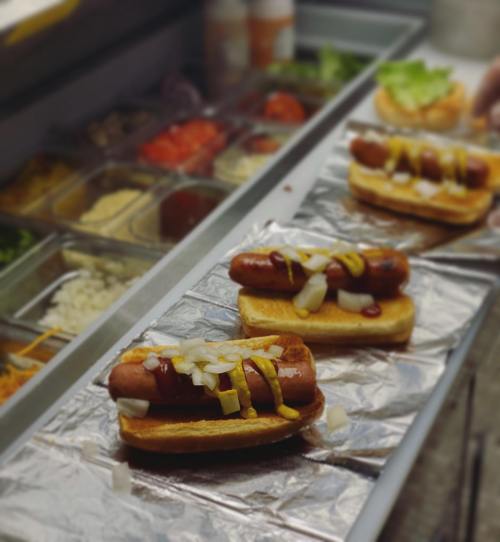 Three hot dogs on buns, topped with mustard, ketchup, and chopped onions, on a counter with various toppings in the background.