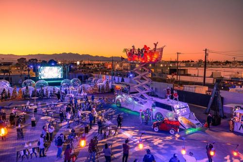 A festive outdoor event at sunset, featuring a decorated vehicle, people mingling, and colorful lights in the background.