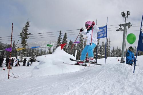 A child in a pink helmet and white jacket skis off a small jump in a snowy landscape, surrounded by colorful flags.