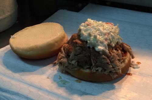 A pulled pork sandwich with coleslaw on a soft bun, resting on a white paper surface.