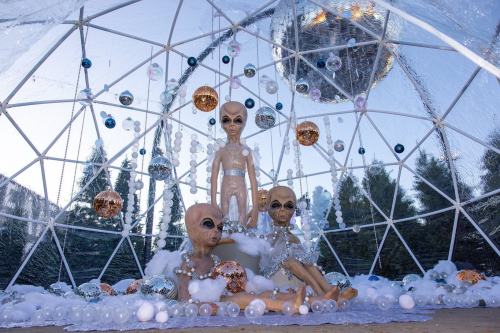 Three alien figures in a decorated dome with ornaments and a disco ball, surrounded by fluffy white clouds.
