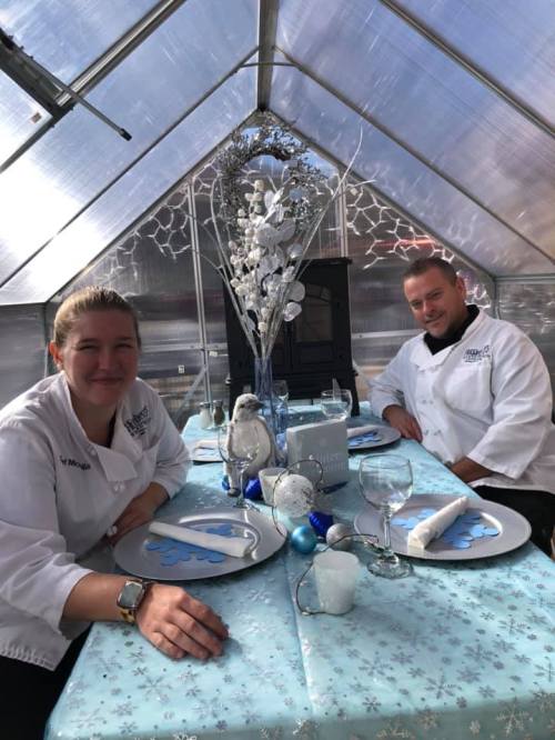Two chefs sit at a beautifully decorated table in a greenhouse, featuring winter-themed decor and elegant table settings.