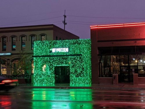 A brightly lit green facade of a building named "Winston," with rain-soaked streets reflecting the lights.