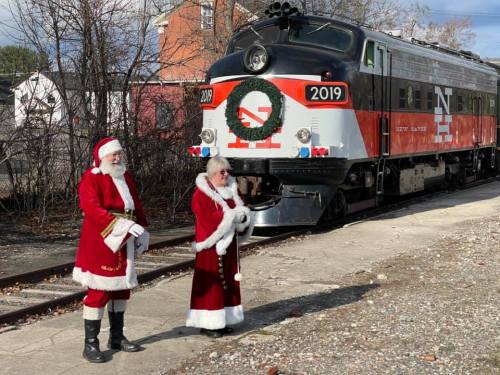 Two people dressed as Santa Claus stand beside a vintage train decorated for the holidays.