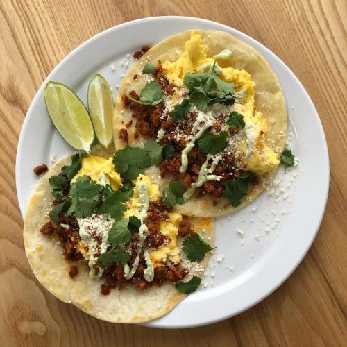 Two tacos filled with scrambled eggs, chorizo, cilantro, and cheese, served with lime wedges on a wooden table.