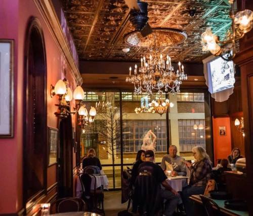 Cozy restaurant interior with chandeliers, ornate decor, and diners enjoying their meals by large windows.