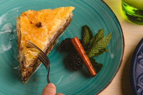 A slice of baklava on a teal plate, garnished with mint, blackberries, and a cinnamon stick, with a fork poised to take a bite.