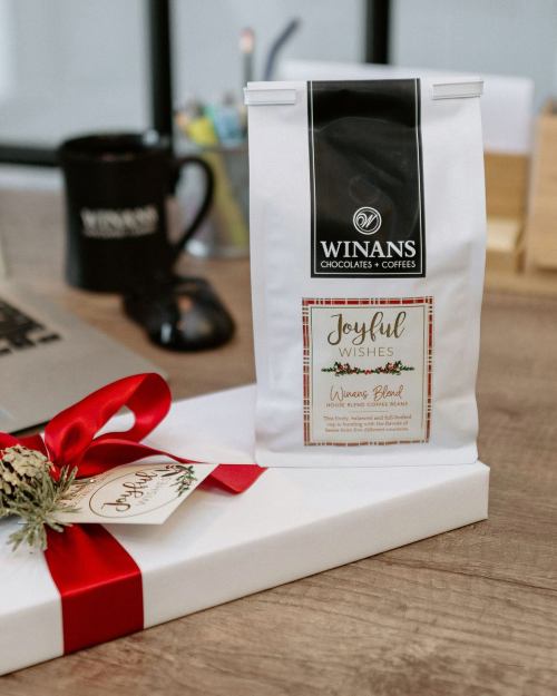 A bag of Winans coffee labeled "Joyful Wishes" sits on a gift box with a red ribbon, next to a coffee mug and laptop.