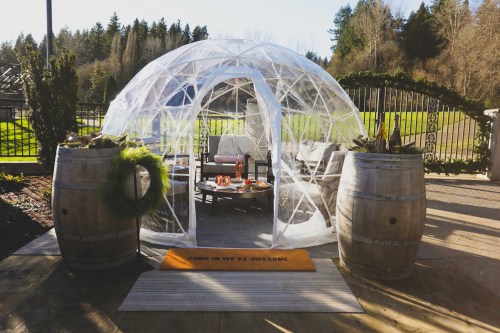 A clear geodesic dome with seating and a welcome mat, surrounded by greenery and wooden barrels.