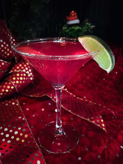 A vibrant pink cocktail in a martini glass, garnished with a lime wedge, set against a festive red background.