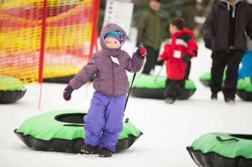 A child in a purple jacket and hat stands on a green snow tube, surrounded by other kids playing in the snow.