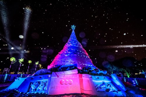 A colorful, illuminated Christmas tree made of sand, surrounded by palm trees and falling snowflakes at night.