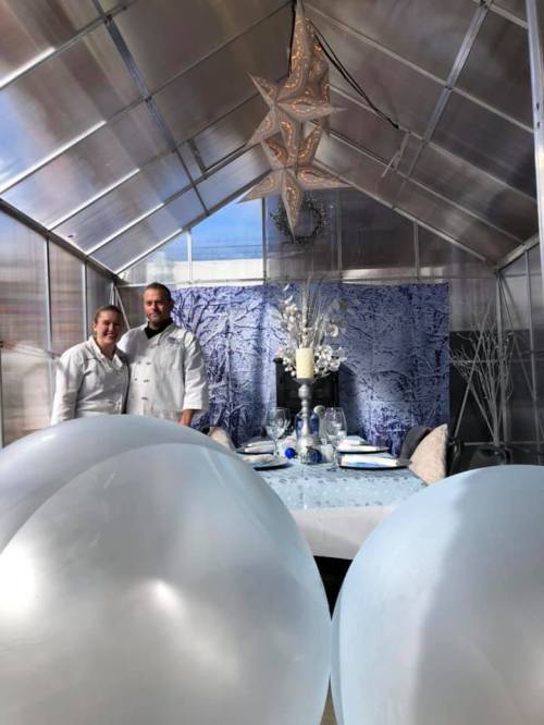 Two chefs stand in a decorated greenhouse with a blue-themed dining setup and large light blue balloons in the foreground.