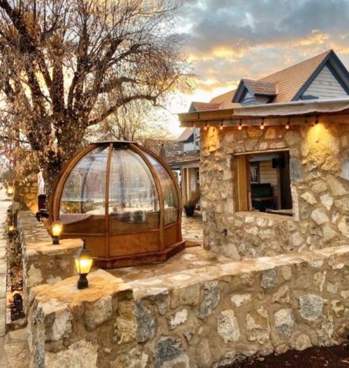 A cozy stone structure with a glass dome, surrounded by trees and warm lighting, under a colorful sunset sky.