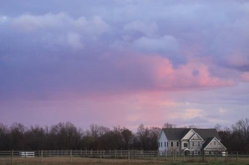 A serene landscape featuring a house beside a fence, with a colorful sunset sky and trees in the background.