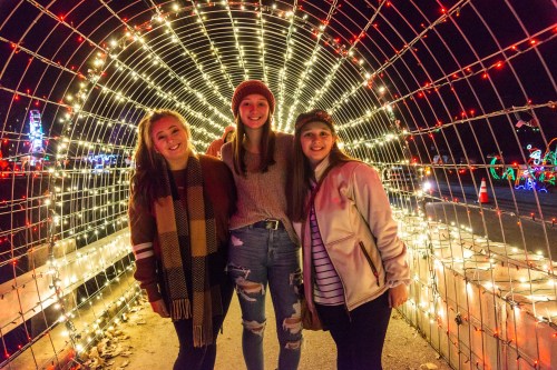 Three friends pose together inside a lit tunnel decorated with colorful holiday lights.