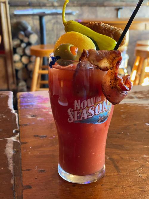 A colorful cocktail garnished with bacon, olives, lemon, and a pepper, served in a glass on a wooden table.