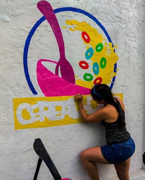 A person paints a colorful cereal-themed mural on a white wall, featuring a spoon and cereal pieces.