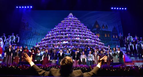 A choir performs in front of a large, decorated Christmas tree on stage, with colorful lights and festive decorations.