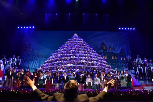A large choir performs on stage in front of a decorated Christmas tree backdrop, with a conductor leading them.