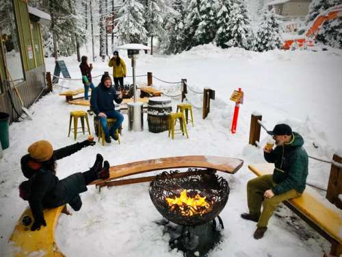 People enjoying drinks around a fire pit in a snowy outdoor setting, with trees and buildings in the background.