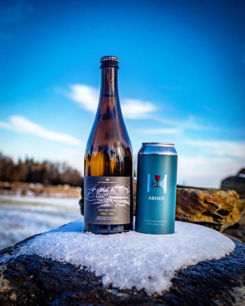 A bottle of wine and a can of beer sit on a snowy rock against a clear blue sky.