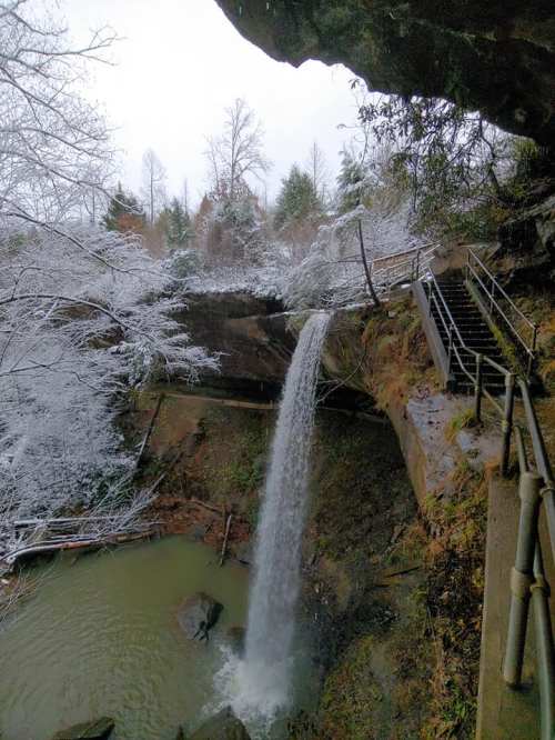 A waterfall cascades into a rocky pool, surrounded by snow-covered trees and a staircase leading down the cliff.