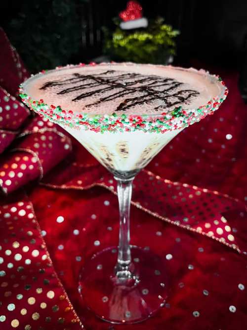 A festive cocktail in a martini glass, rimmed with colorful sprinkles, on a red and gold polka dot background.