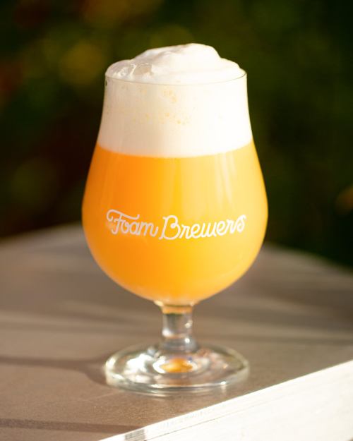 A glass of hazy yellow beer with a frothy white head, featuring the logo "Foam Brewers" on the side.